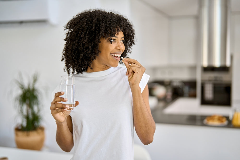 Woman in a kitchen holding a glass of water and a supplement