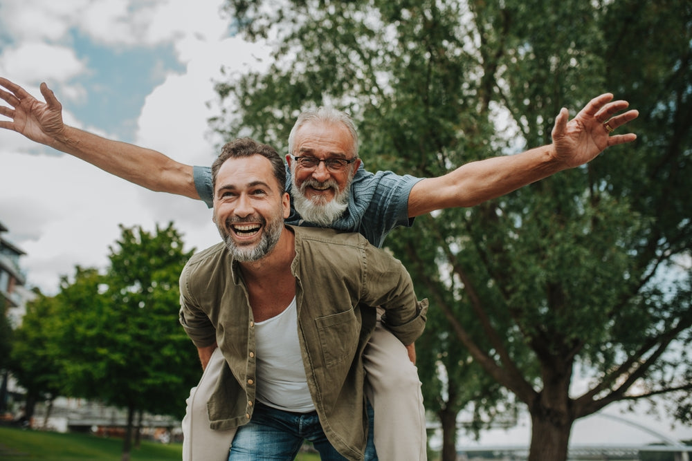 Two men enjoying a piggyback ride in a park with trees and blue sky in the background.